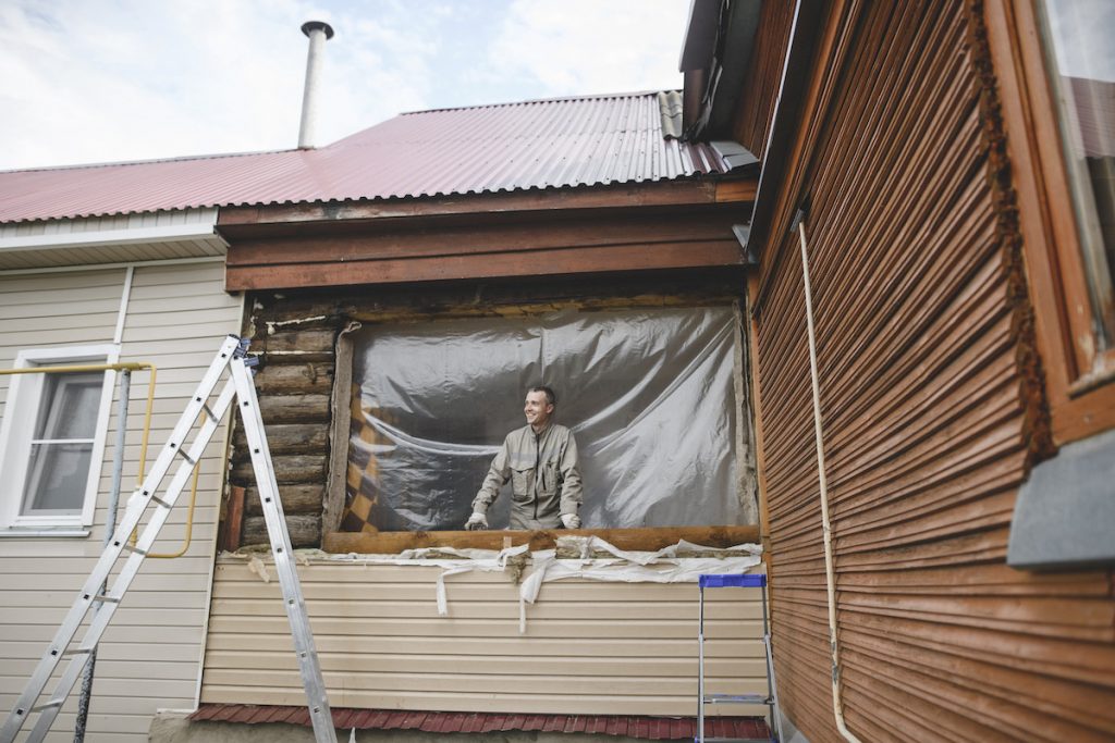 A man peering outside of his home from a large space on the home's siding where a window will be installed.