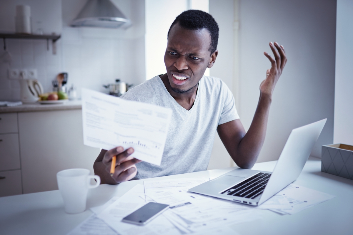 Man sitting in front of his laptop looking at a piece of paper in his hand angrily.