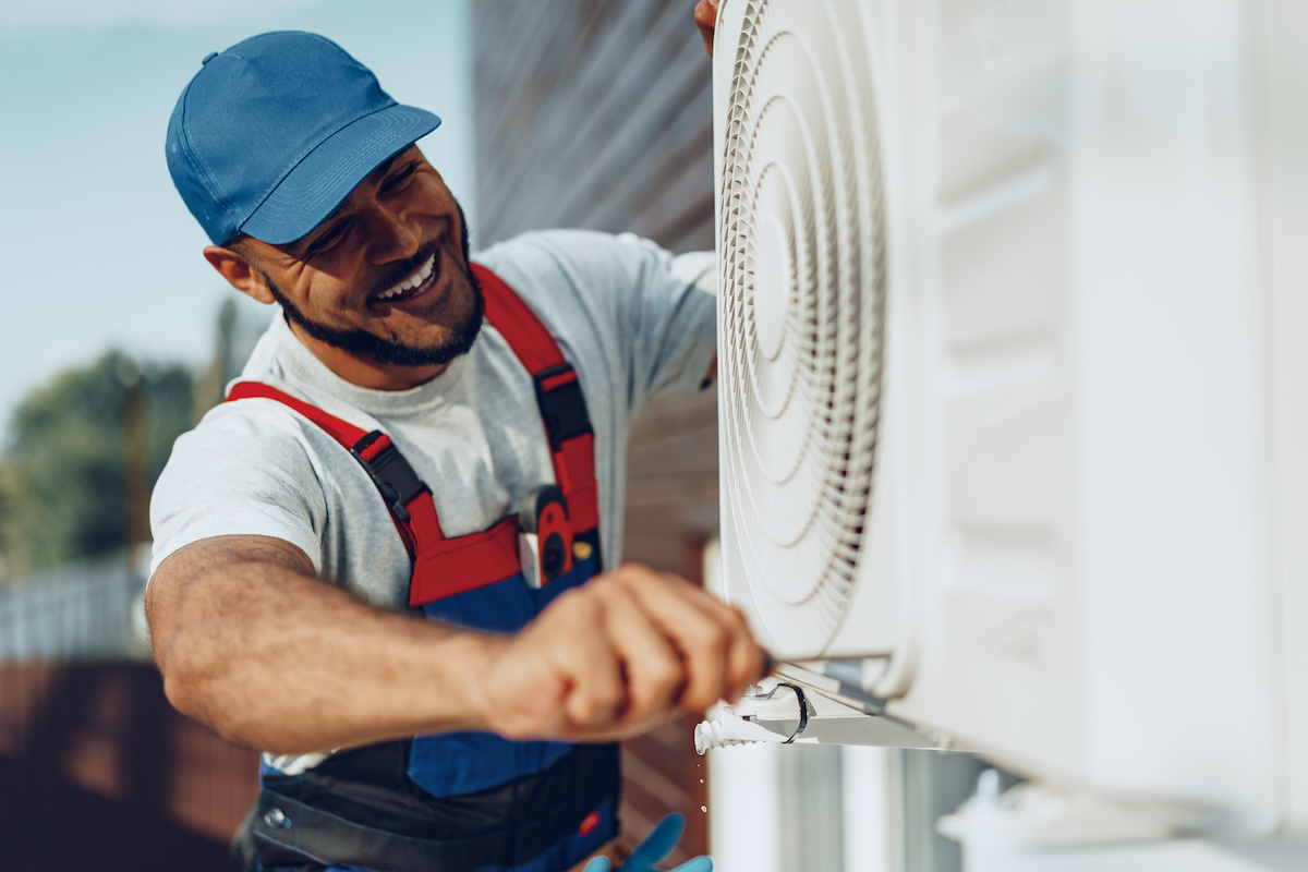 Man wearing a blue hat and overalls while installing a new AC unit and smiling. Average cost to replace ac unit in Florida.