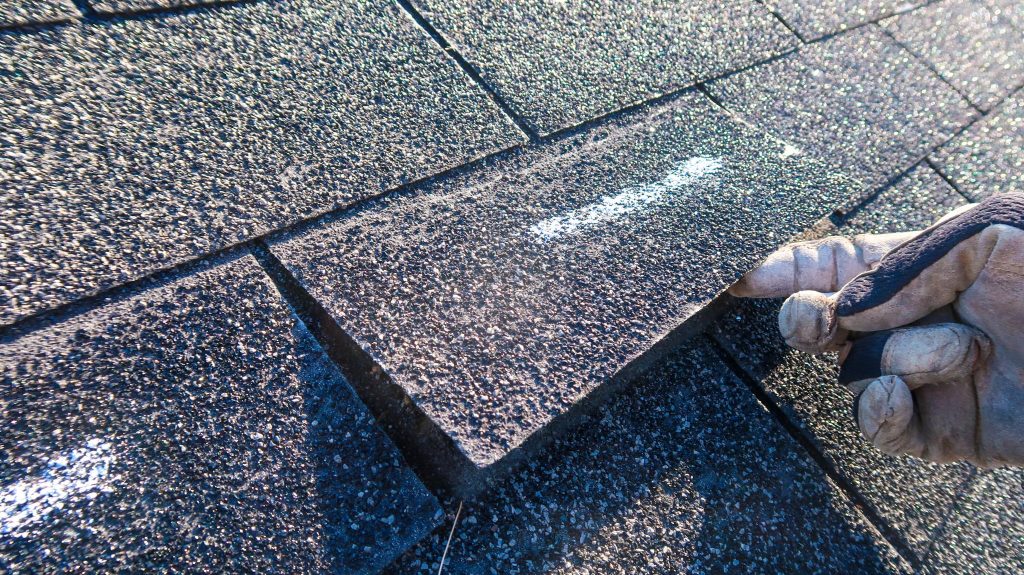 Close-up of a hand wearing a work glove, installing a new black asphalt shingle on a roof, with a focus on the texture of the shingle.