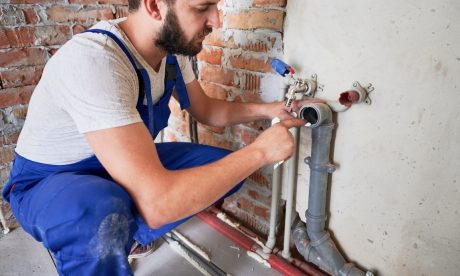 A plumber wearing a white hard hat and blue overalls works on a sewer pipe attached to a brick wall in an unfinished building.