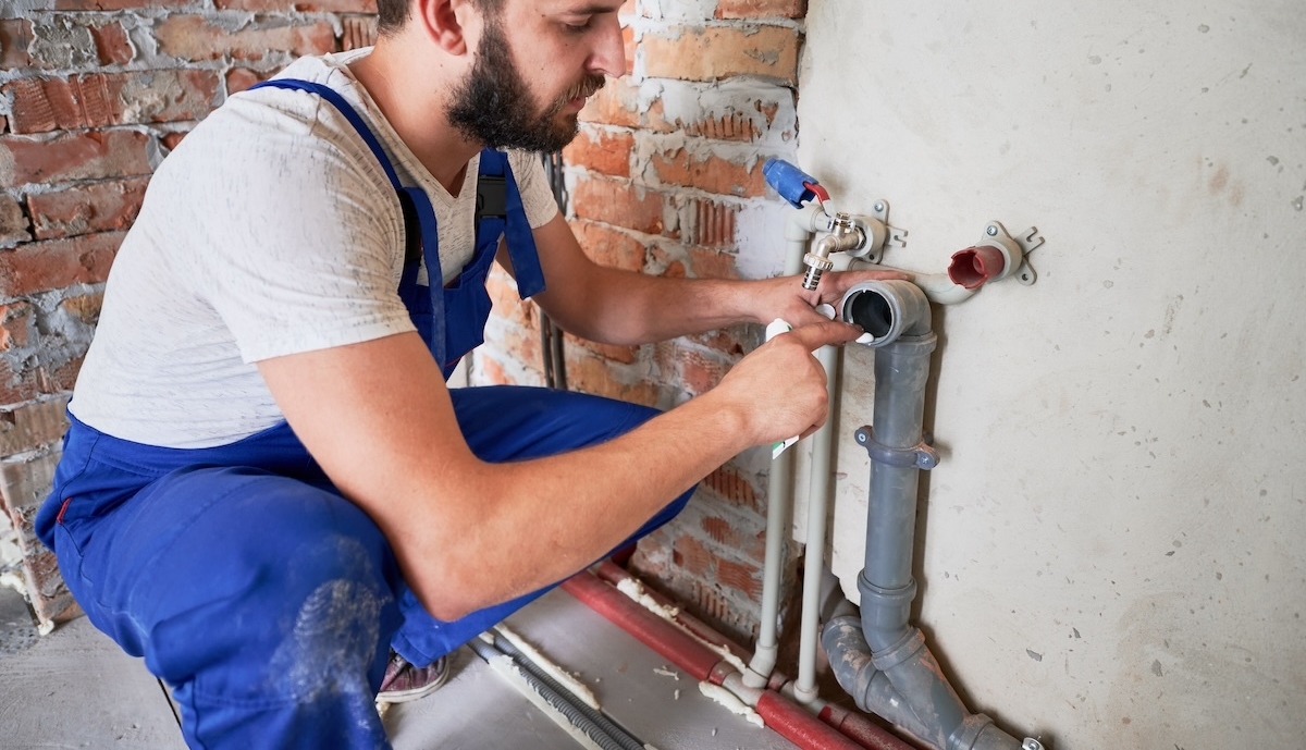 A plumber wearing a white hard hat and blue overalls works on a sewer pipe attached to a brick wall in an unfinished building.