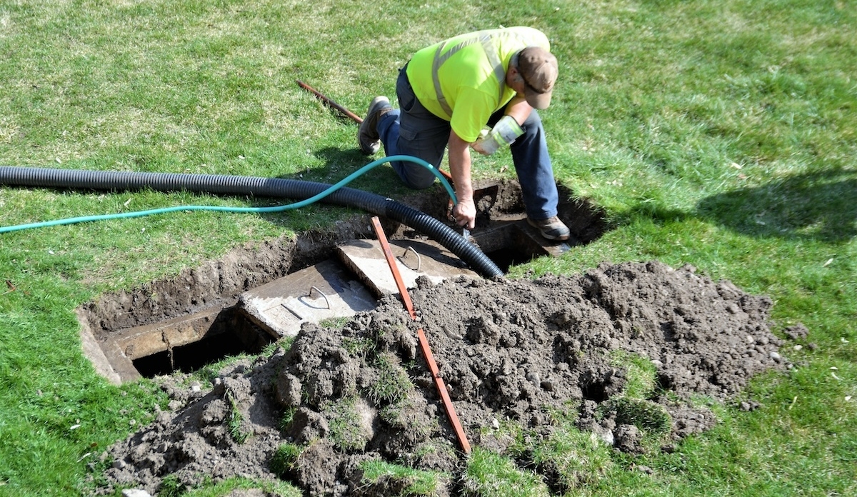 A worker in a neon yellow safety shirt and brown cap kneels on a lawn, installing a septic drain pipe into an underground system.