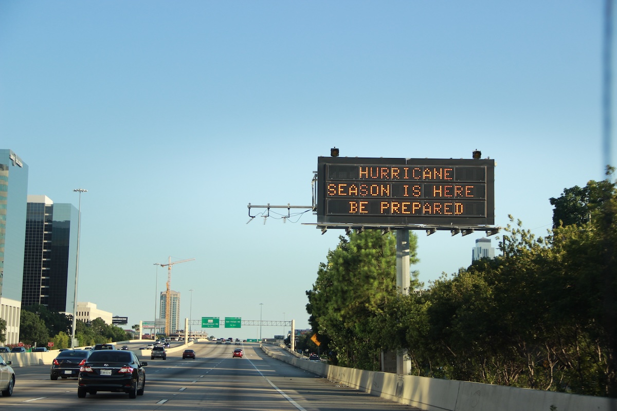 Highway digital sign reads "Hurricane Season is Here. Be Prepared" with cars and city buildings in the background.