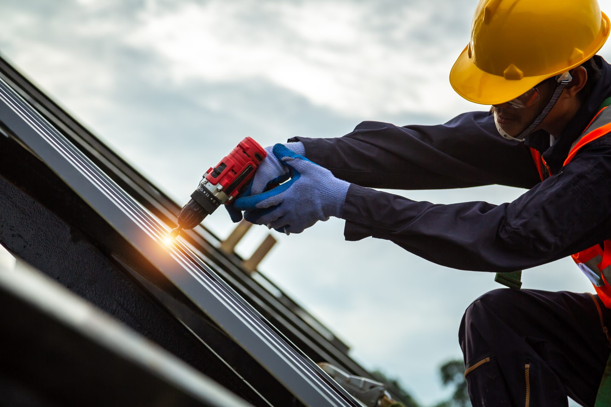 A construction worker in a yellow hard hat and safety gear uses a red power drill on a metal surface, with sparks flying where the drill makes contact.