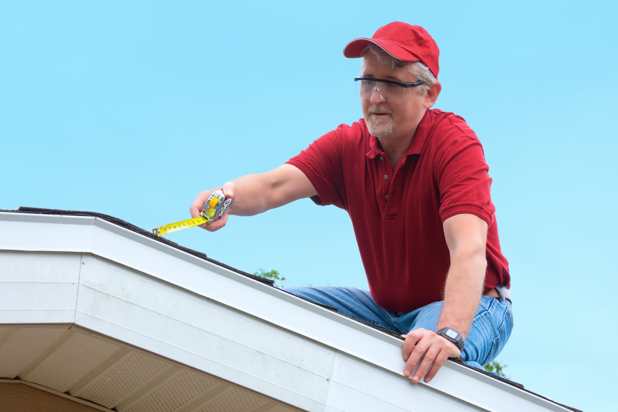 A man in a red shirt and cap uses a tape measure to inspect the edge of a roof.