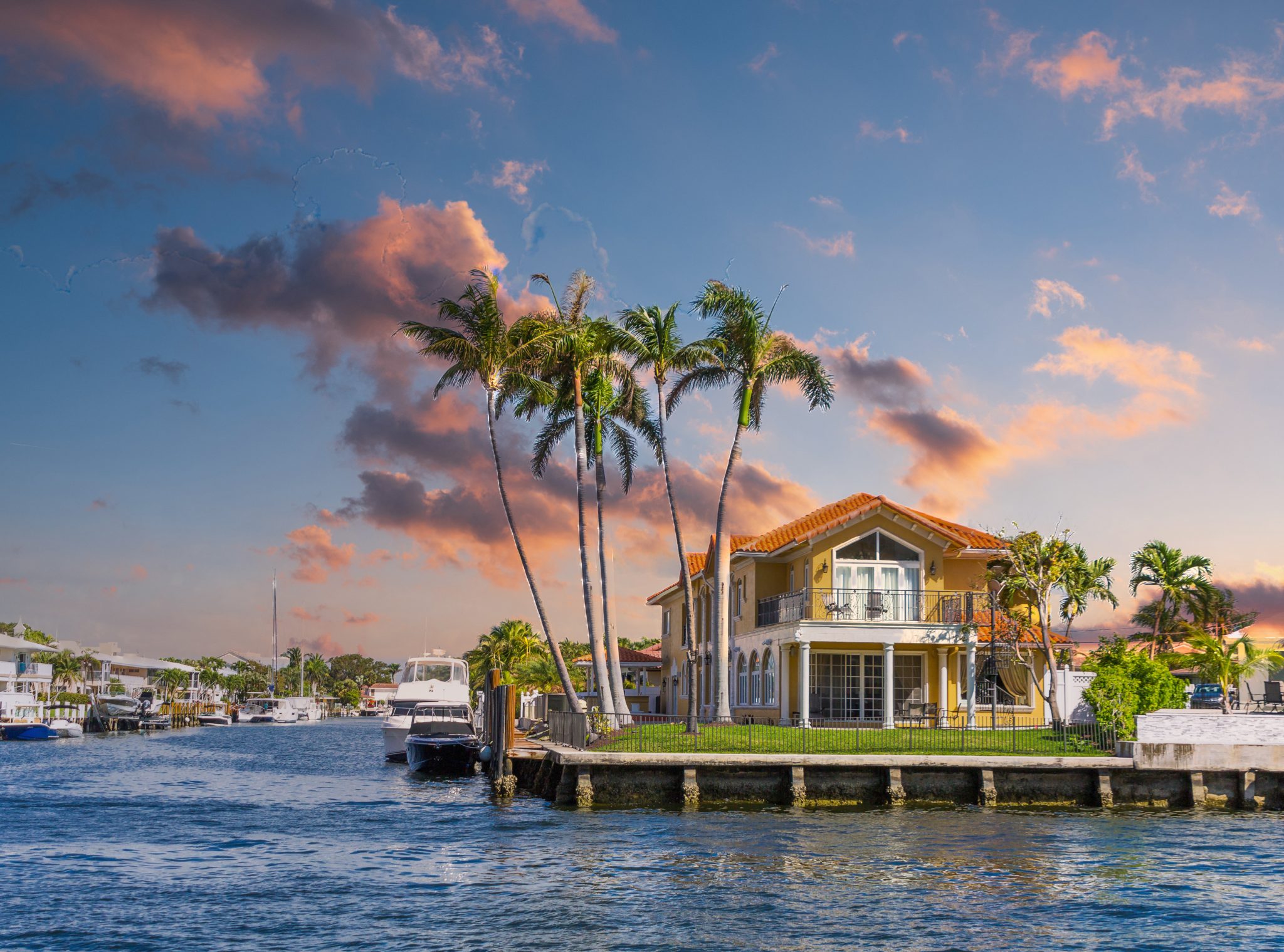 A luxurious Florida house by the water featuring palm trees and a dock with boats under a vibrant sunset.