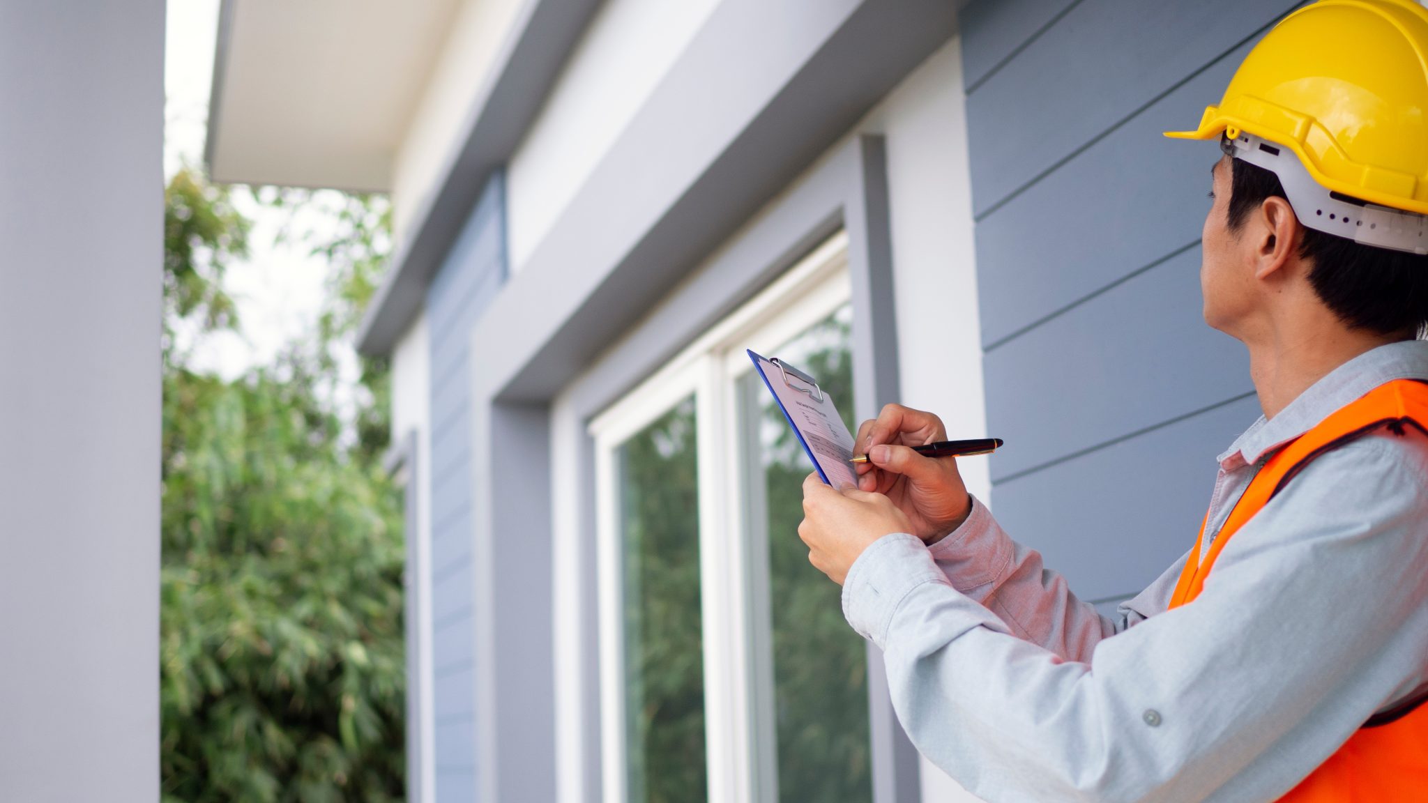 A construction worker in a yellow hard hat and orange safety vest inspects the exterior of a house, taking notes on a clipboard.