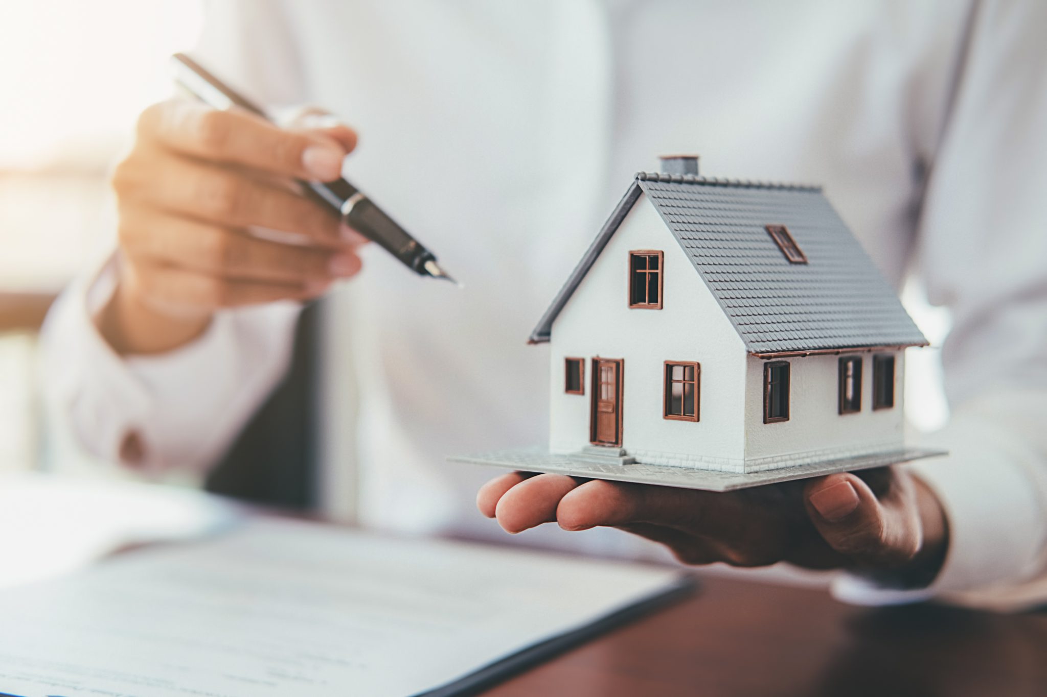 Close-up of a person’s hand holding a miniature house and a pen, with a document on the table, indicating a real estate or insurance transaction.