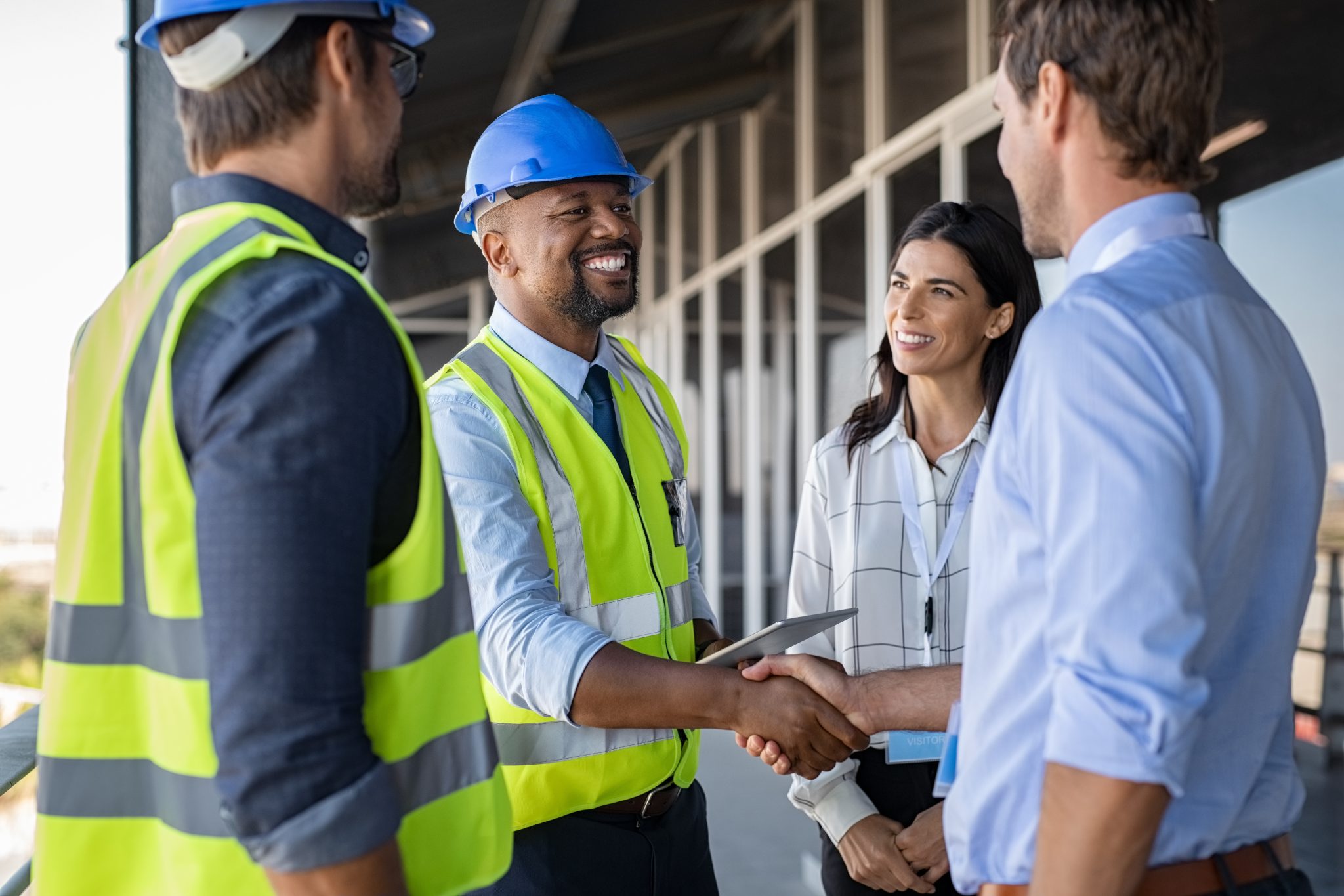 A smiling construction worker in a blue hard hat and yellow safety vest shakes hands with a man in business attire at a construction site, while another worker and a woman look on with smiles.