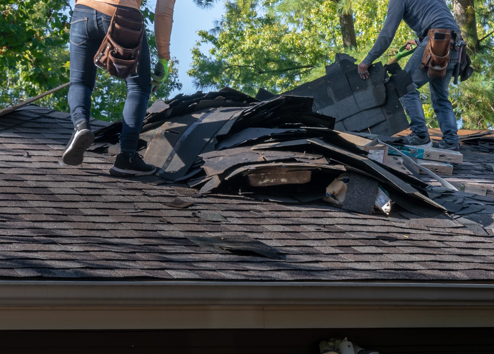 Two workers on a roof removing damaged shingles and debris as part of post-storm repair, wearing gloves and protective gear.