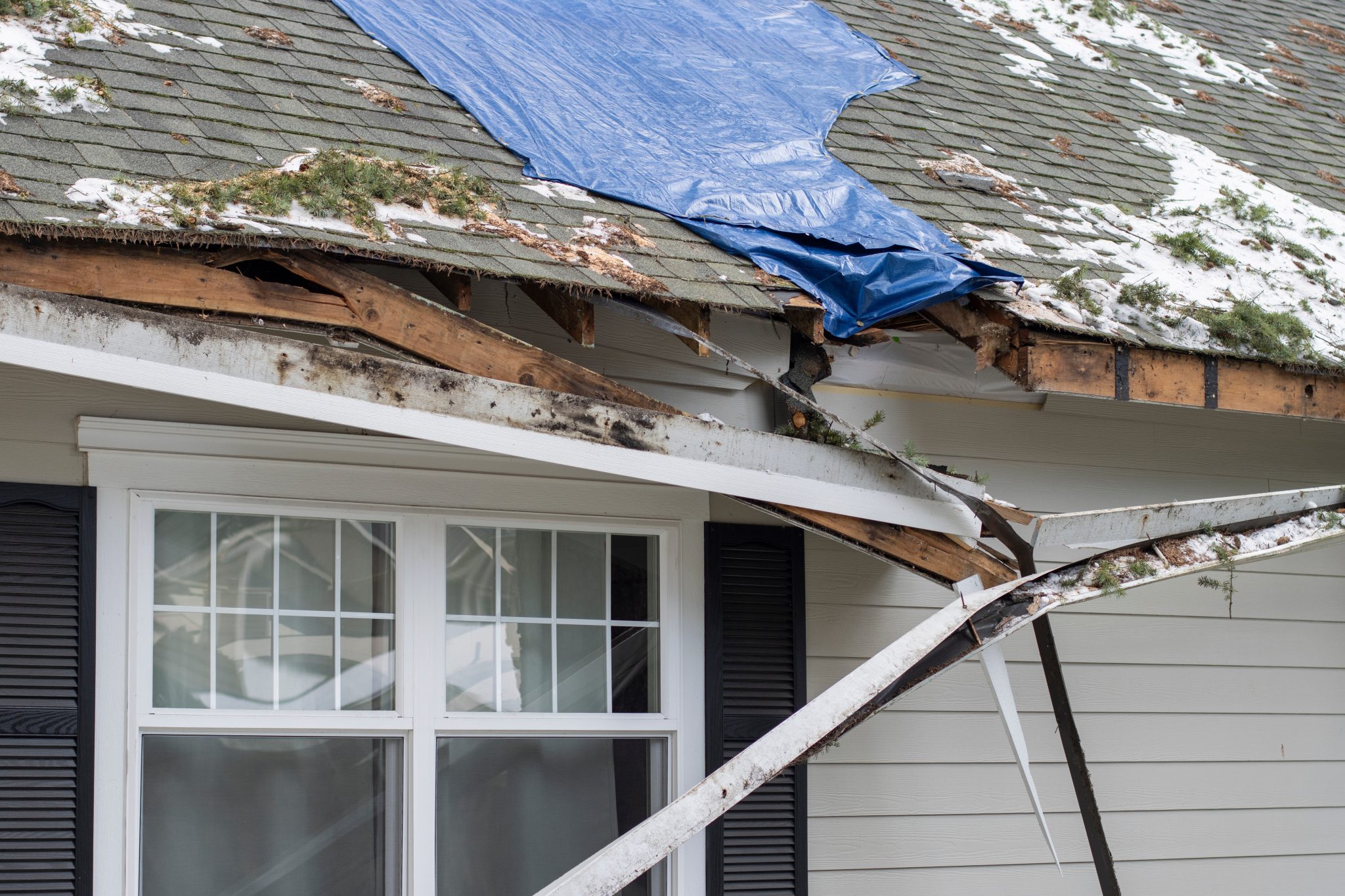 Damaged roof with a blue tarp covering a section of missing shingles and exposed wood, showing structural impact from storm damage.