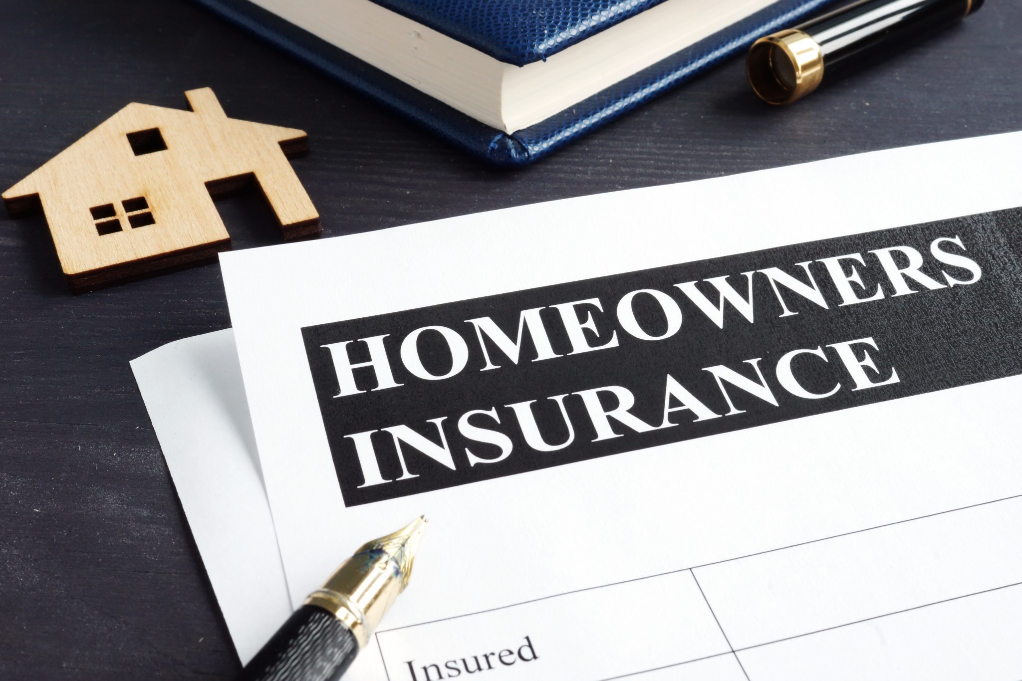 "Homeowners Insurance" document on a dark desk, accompanied by a wooden house figurine, a blue book, and a pen, symbolizing property protection and financial planning.