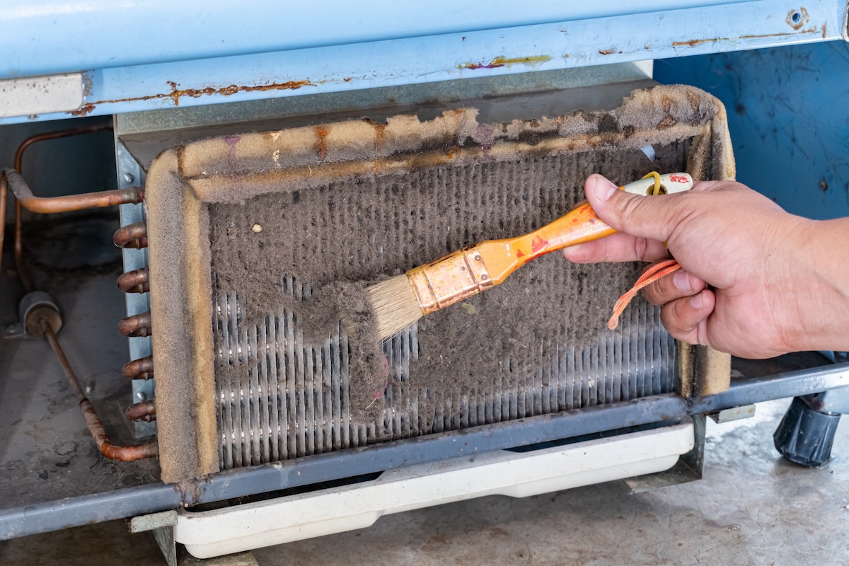 A man's hand using a brush to clean the dirty condensing unit on a home HVAC system.