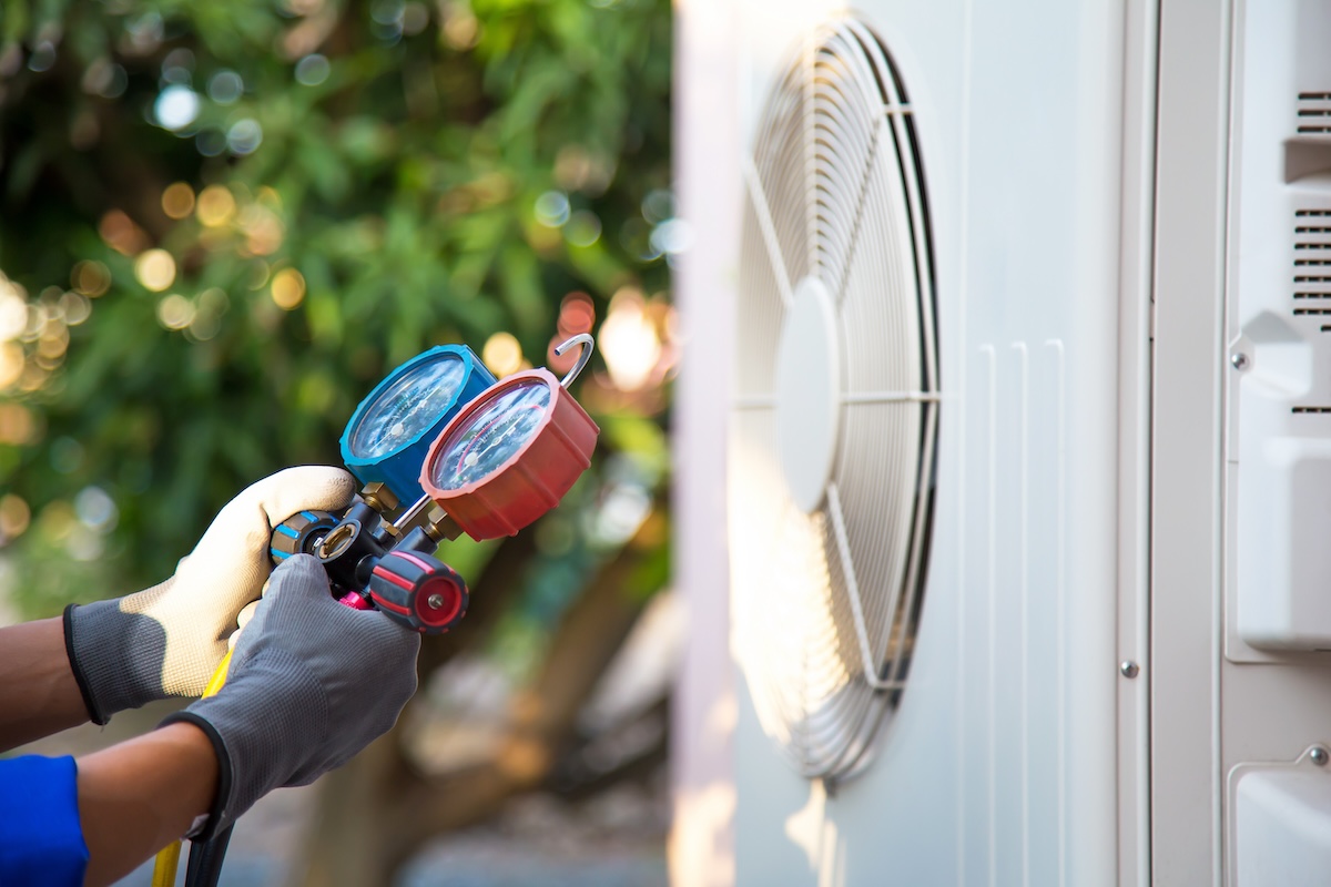 A technician wearing gloves holds a manifold gauge set while servicing an outdoor air conditioning unit.